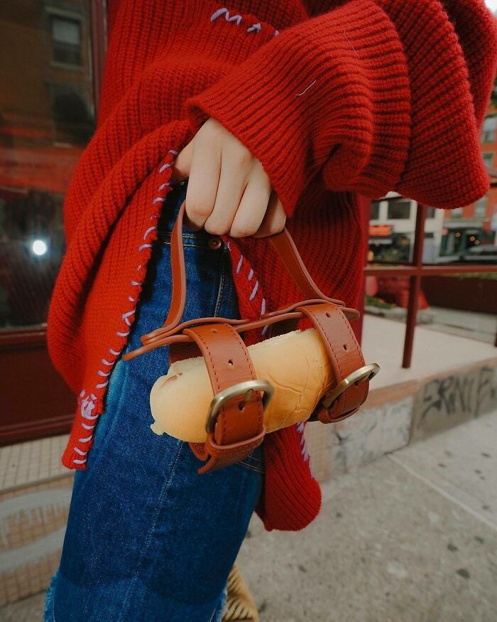 Person wearing a red sweater holding an unusual accessory by Nik Bentel featuring a bread roll with leather straps as a handbag.