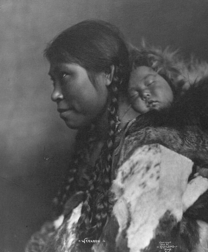 Historic black and white photo of an Inuit mother carrying her sleeping child wrapped in fur clothing.