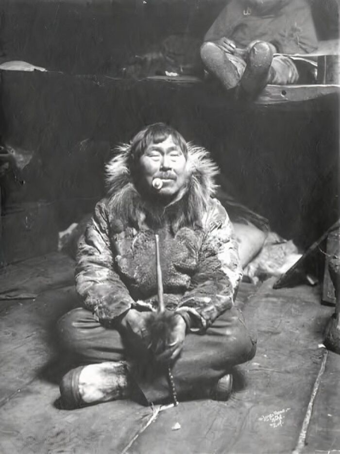 Inuit man in traditional fur clothing sitting cross-legged indoors, holding a tool, historic photo capturing Inuit spirit.