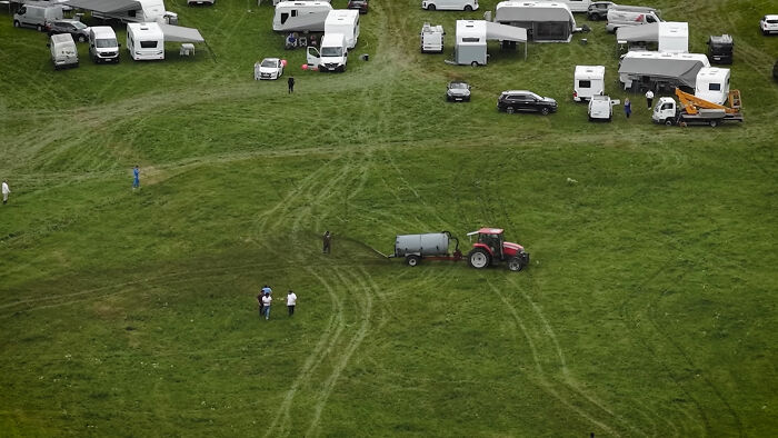 Aerial view of a farm vehicle spraying manure near camper vans and people on a grassy field.