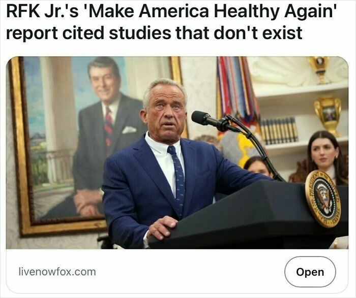 Man in suit speaking at presidential podium with painting and people in background, satire headlines topic.