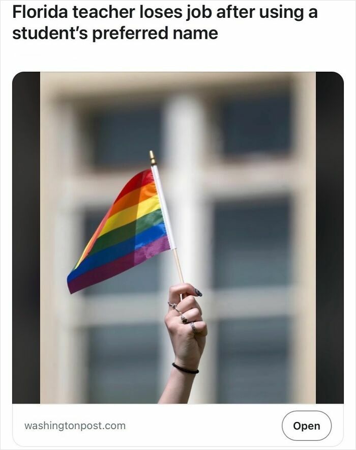 Hand holding a small rainbow flag against a blurred building background representing headlines that seem like satire but are true.