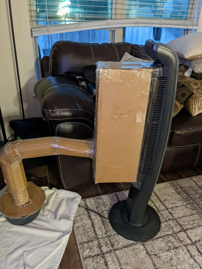 Ventilador con sistema casero de cartón para enfriar durante olas de calor veraniegas en sala con sofá y ventana.