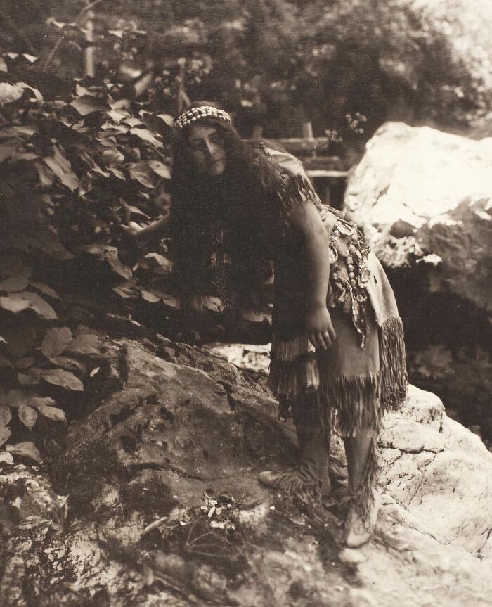 Native tribes member in traditional attire standing on rocky terrain surrounded by foliage, early 1900s strength captured.