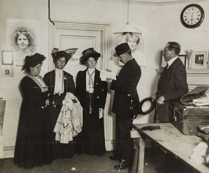 Women in the 20th century suffrage era dressed in period clothing, interacting with a police officer indoors.