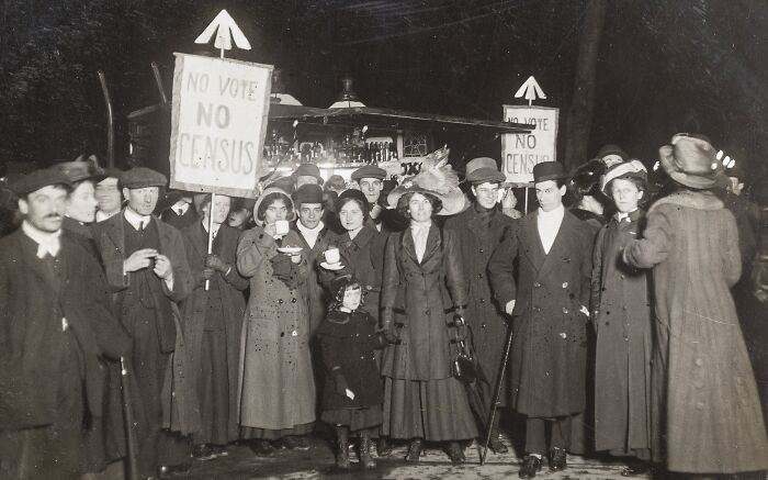 Group of women and men in 20th century suffrage era protesting with signs demanding voting rights.
