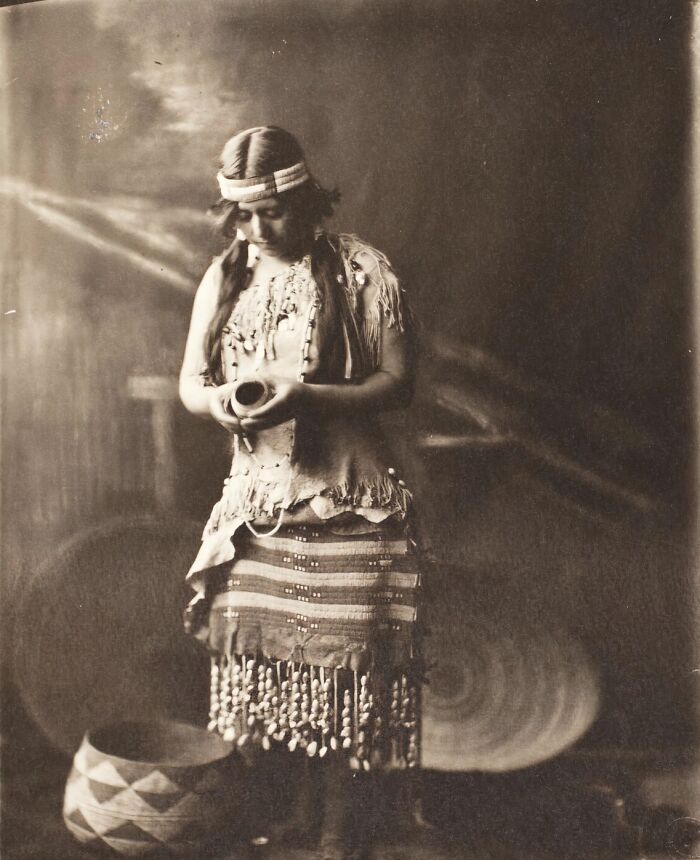 Native tribes woman in traditional dress holding a pottery vessel with woven baskets in the background, 1900s.