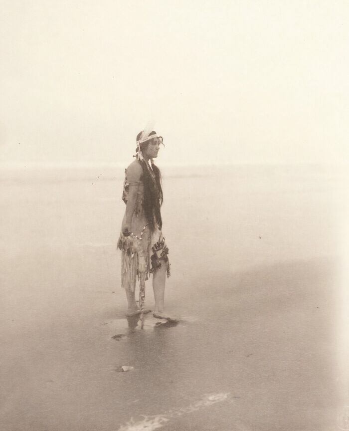Native tribes member in traditional attire standing barefoot in shallow water, captured in early 1900s photography.
