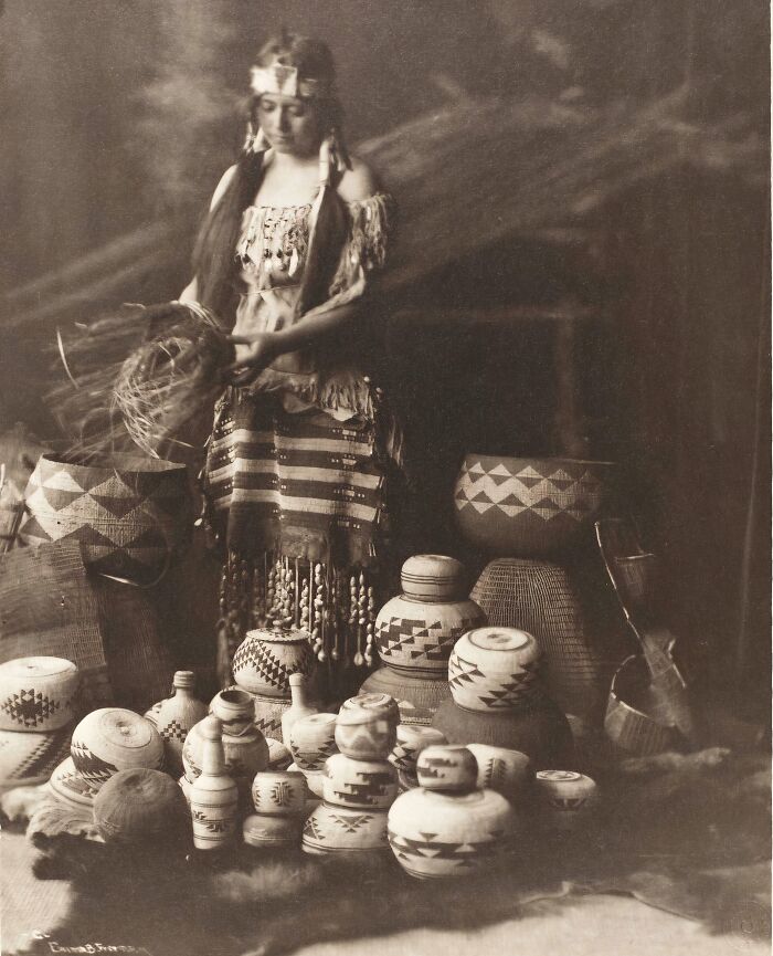 Native tribes woman in traditional dress surrounded by woven baskets showcasing strength and culture in early 1900s photography.