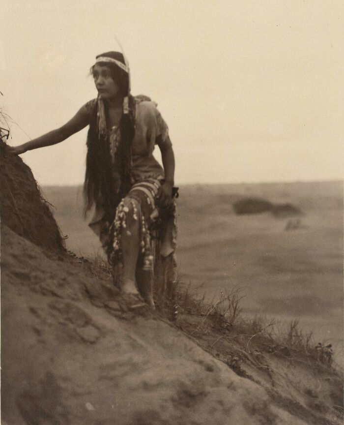 Native tribes woman in traditional dress climbing a hillside, historical black and white photo capturing strength and culture.