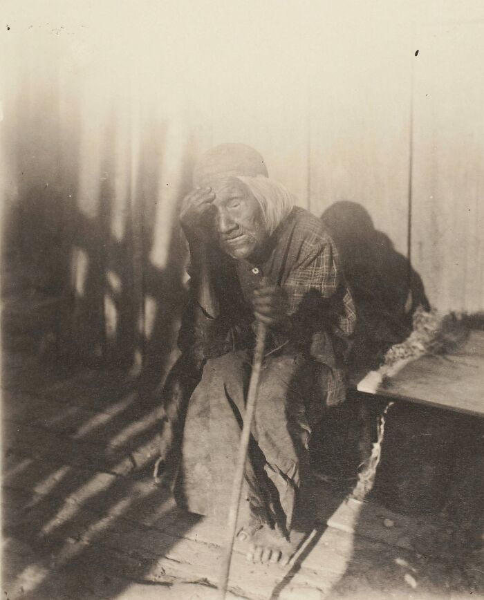 Elderly Native tribesperson in traditional clothing sitting with a staff, captured in a vintage black and white photo.