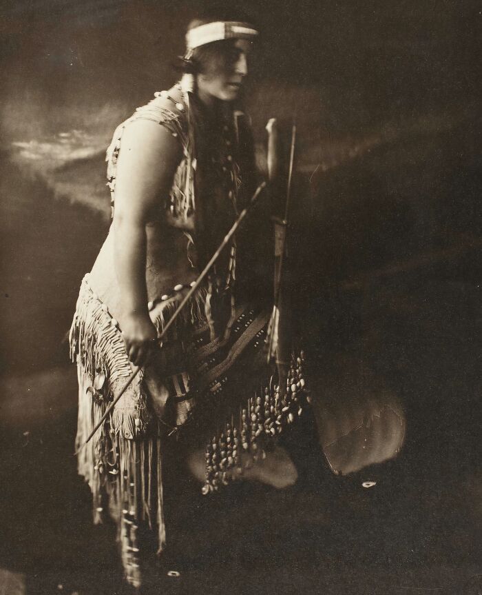 Vintage photo of a Native tribes member in traditional attire holding cultural artifacts, showcasing strength and heritage.