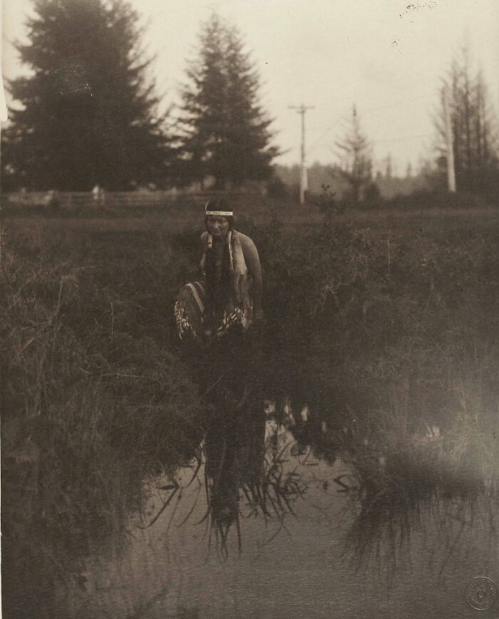 Native tribes member in traditional attire by a pond in a wooded area, captured in a historic early 1900s photograph.