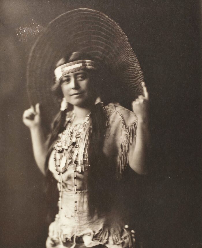 Native tribes woman wearing traditional clothing and large hat, captured in a vintage black and white photograph from the 1900s.