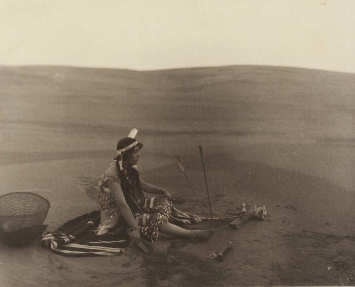 Native tribes member in traditional attire sitting on a blanket with spear and basket in a desert, capturing strength in 1900s.