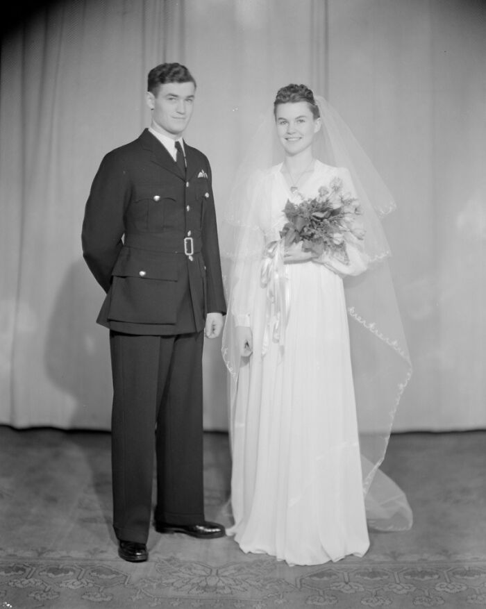 Black and white 1940s wedding photo of a bride in a veil holding flowers and a groom in military uniform smiling indoors.