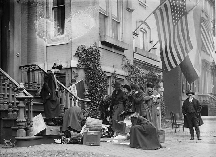 Group of women in early 20th century clothing gathering documents on a city street during the suffrage era.