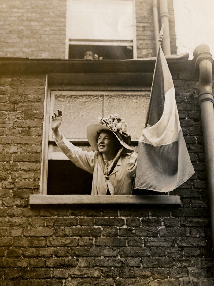 Woman in 20th century suffrage era attire waving from a window next to a flag on a brick building wall.