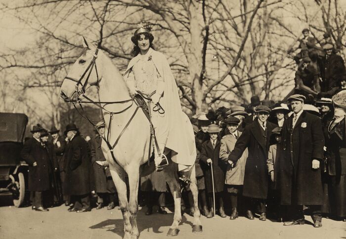 Woman in suffrage era attire riding a white horse during a 20th century women's rights public gathering.