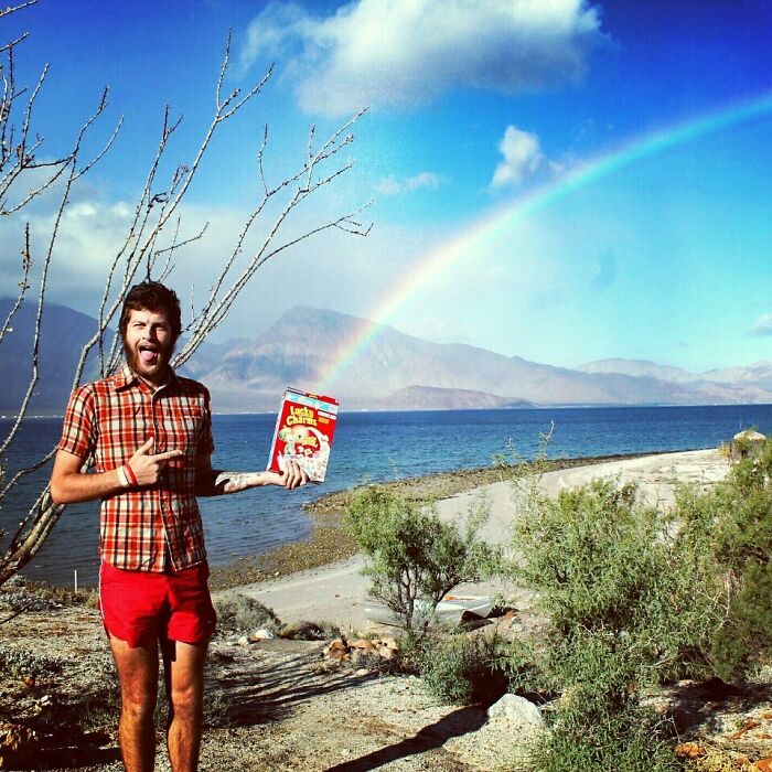 Man in red shorts and plaid shirt holding a cereal box by a lake with a rainbow, a funny vacation photo moment