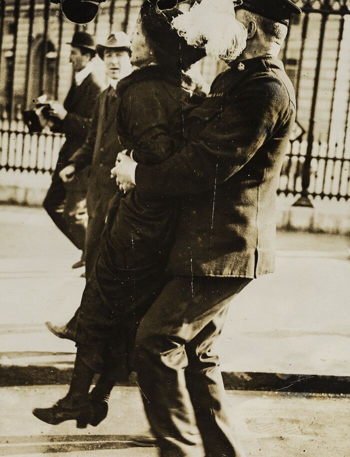 Woman in 20th century suffrage era attire being carried by a uniformed officer during a protest or event.