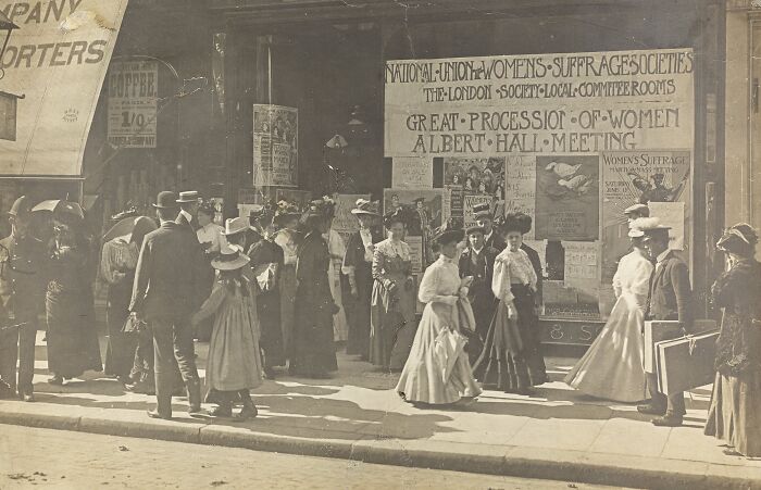 Crowd of women in early 20th century clothing gathering outside building with women's suffrage era protest signs and banners.