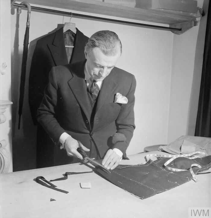 Tailor cutting fabric with scissors in a workshop, showcasing British women’s style adaptations during WWII restrictions.