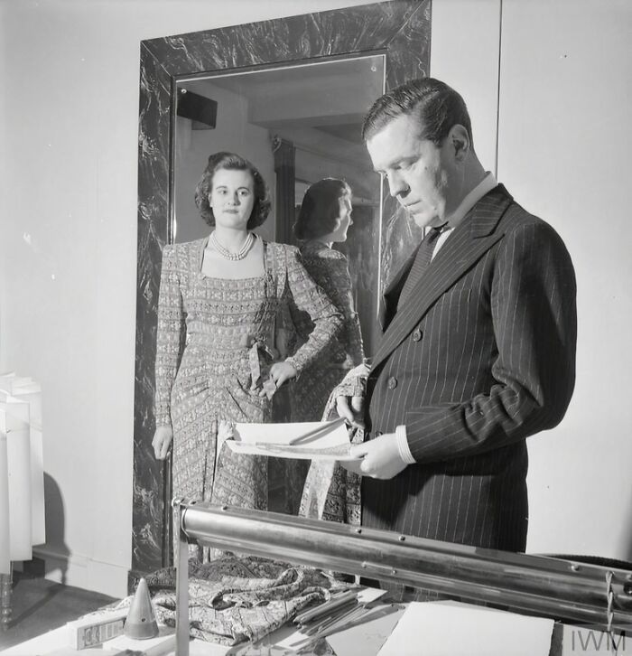 British woman dressed in WWII era style dress standing near a man reviewing documents inside a room.