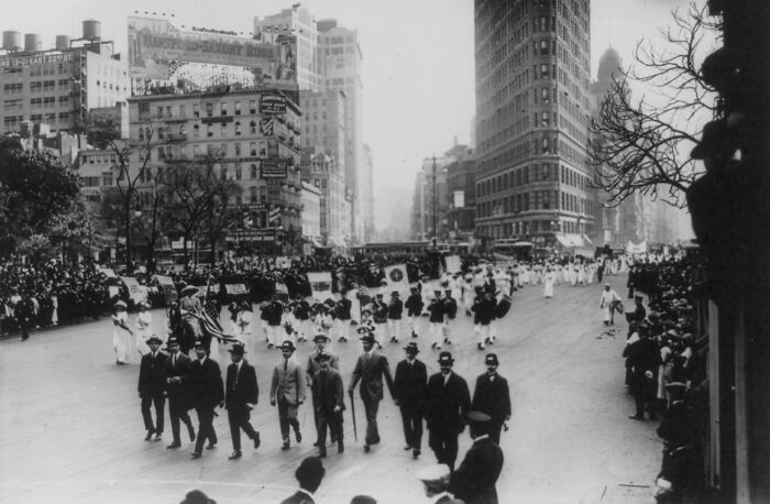 Crowd and women marching in a 20th century suffrage era parade on a city street with tall buildings and spectators.