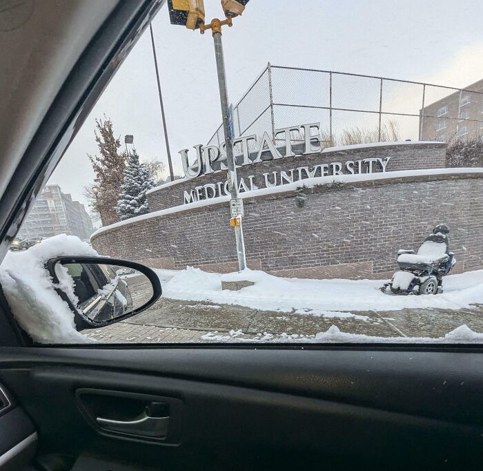 Snow-covered street scene outside Upstate Medical University, showing a wheelchair and traffic signal in winter weather.