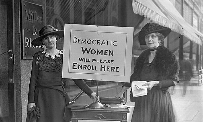 Two women in early 20th century clothing standing by a sign encouraging democratic women to enroll during the suffrage era