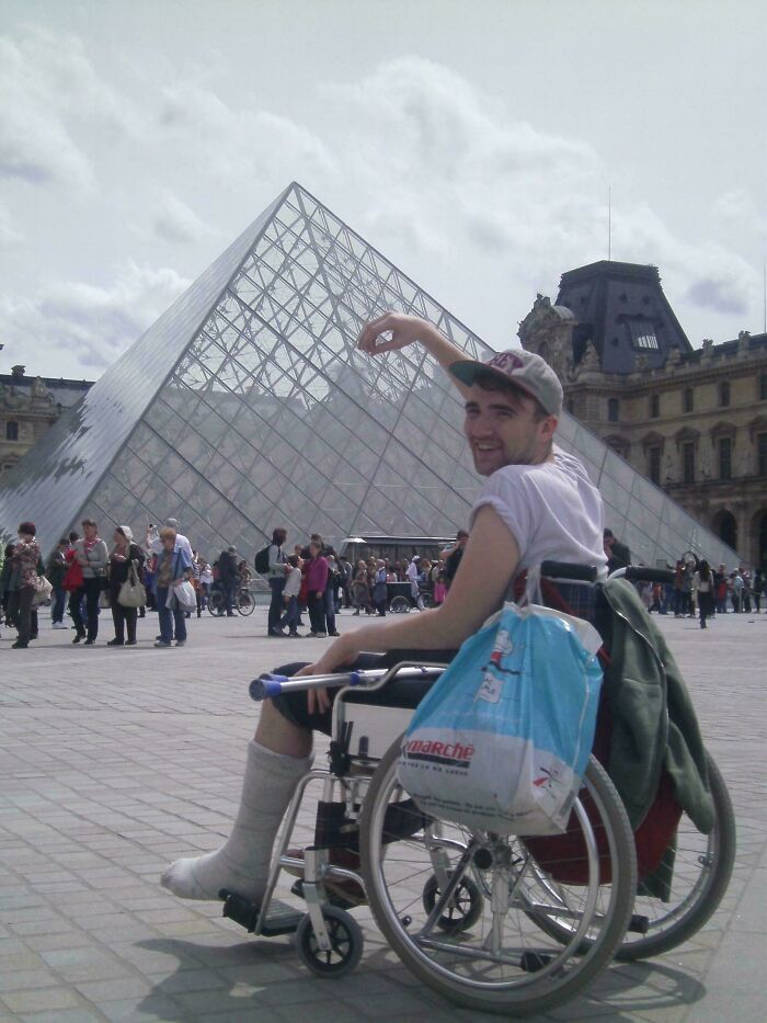 Young man in wheelchair with a cast on his leg posing for a funny vacation pic at the Louvre Pyramid in Paris.