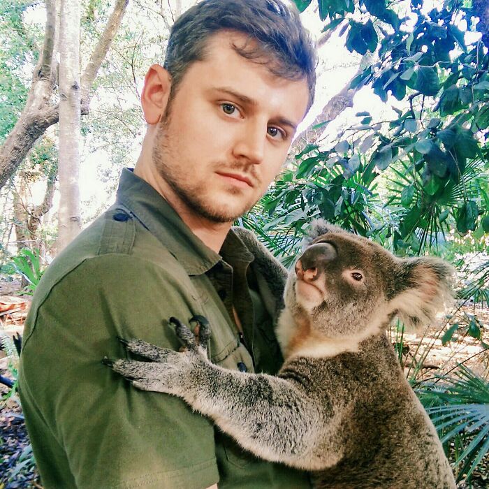 Man holding koala during a vacation pic in a lush forest setting, capturing a funny travel moment.