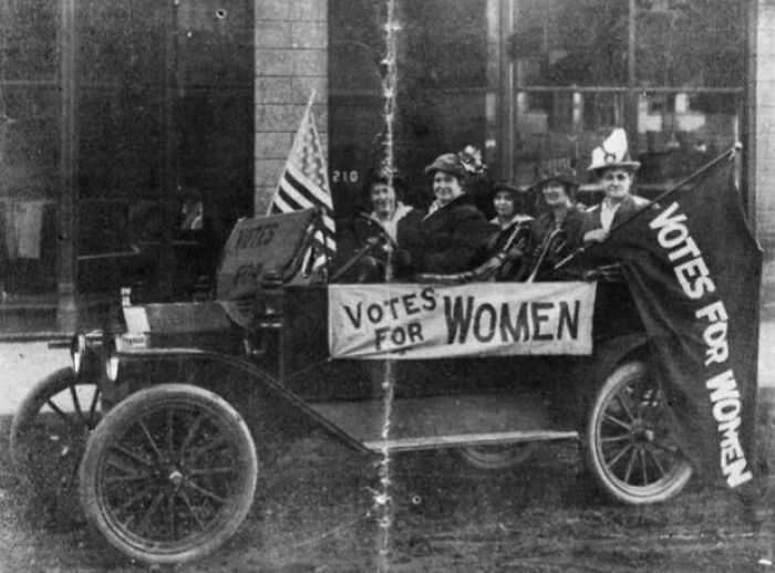 Women in the 20th century suffrage era riding in a car with Votes for Women banners and an American flag.