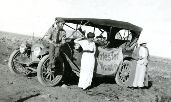 Women in 20th century suffrage era standing by early automobile with votes for women banner in rural area.