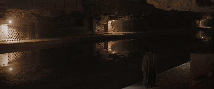 Dimly lit ancient stone structure with reflective water and a cloaked figure, highlighting bad movie details in a mysterious scene.