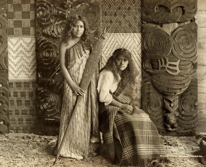 Two Māori girls in traditional clothing posed with carved wooden panels in a powerful portrait of Māori life.
