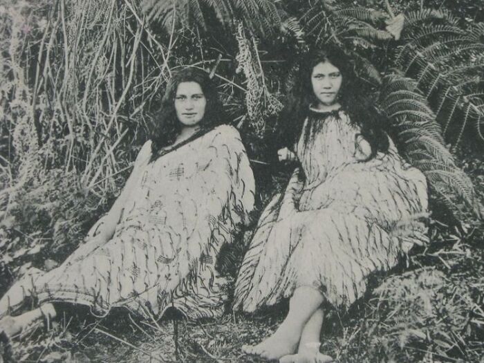 Two Māori women wearing traditional cloaks, sitting outdoors surrounded by native New Zealand foliage in a historic portrait.