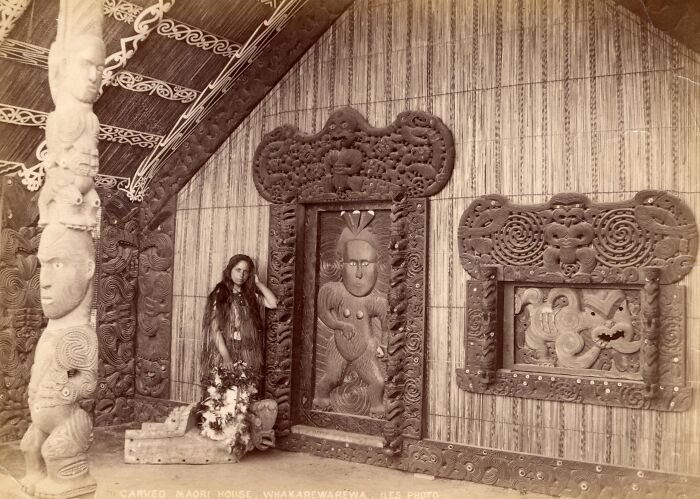 Woman standing inside a carved Māori house surrounded by intricate woodwork and traditional Māori art from a century ago