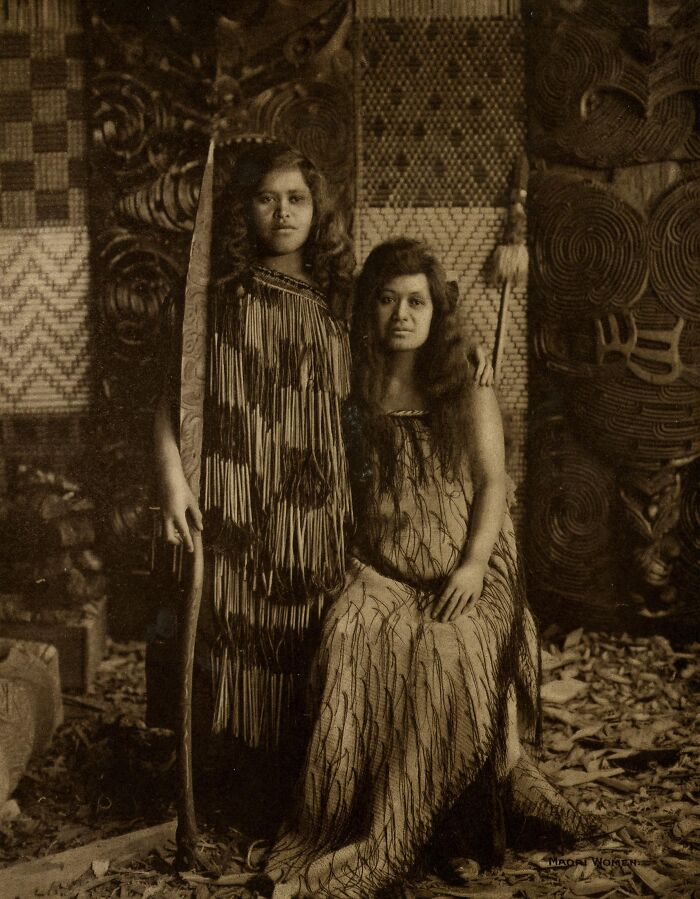 Two Māori women in traditional dress posing indoors surrounded by woven textiles, representing Māori life a century ago.