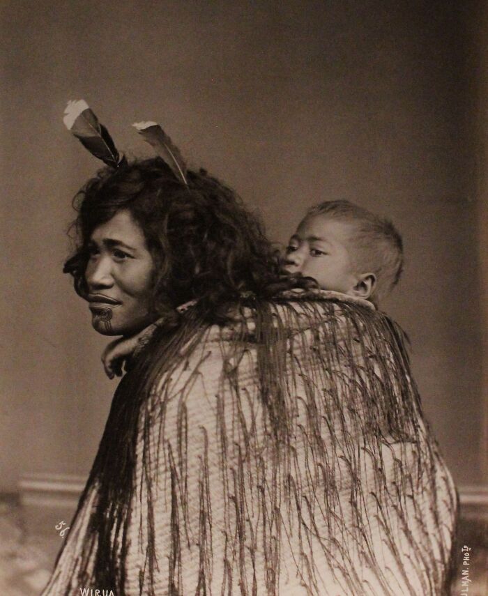 Māori woman with traditional facial tattoo and feathers carrying a child wrapped in a woven cloak in a powerful portrait.