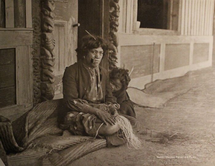Māori woman twisting flax with child sitting beside her in a traditional setting, showcasing Māori life a century ago.