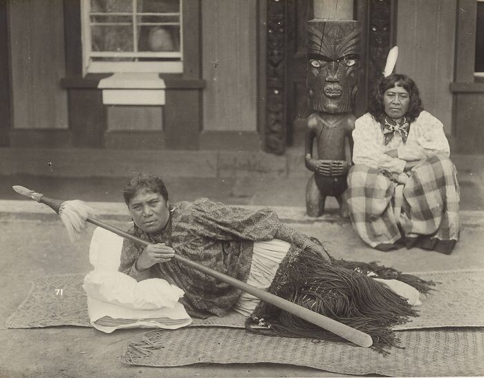 Two Māori women in traditional dress pose with a carved wooden figure, capturing Māori life a century ago.