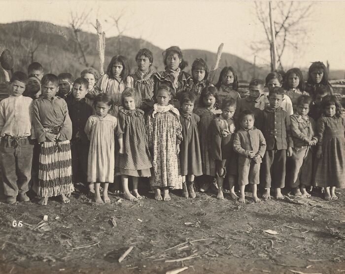 Group portrait of Māori children standing outdoors in traditional and early 20th-century clothing capturing Māori life a century ago