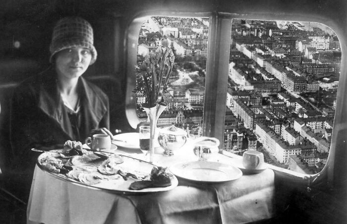 Woman dining elegantly by airplane window overlooking cityscape, showcasing the glamour of flying in the past.