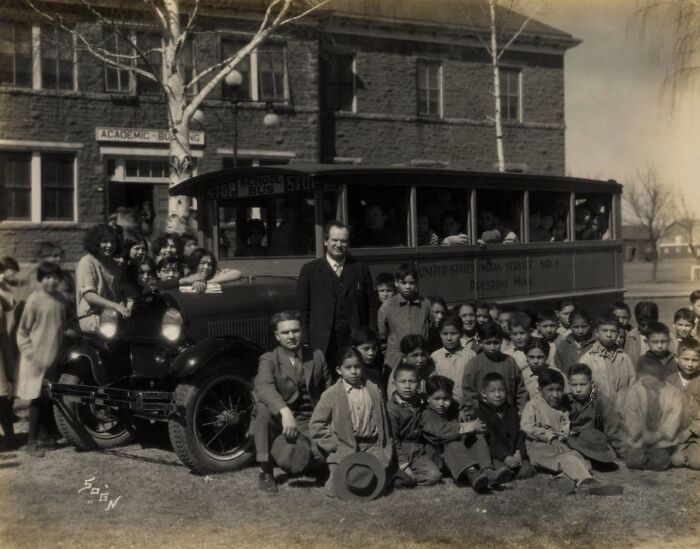 Group of Native American children and staff posing with a school bus outside a boarding school in the 1910s.