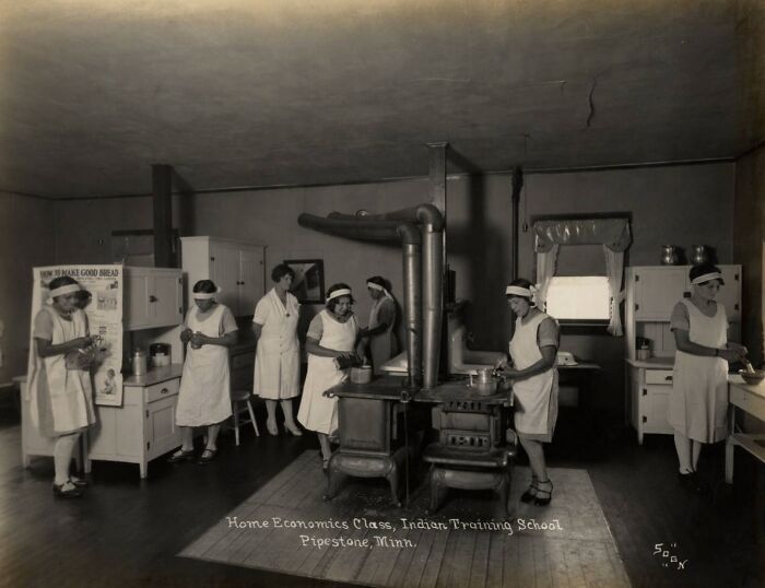 Native American girls in 1910s boarding school home economics class preparing food around a stove in a classroom.