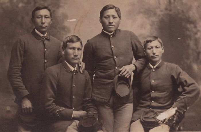 Four Native American boarding school students in 1910s uniforms posing for a formal studio portrait.