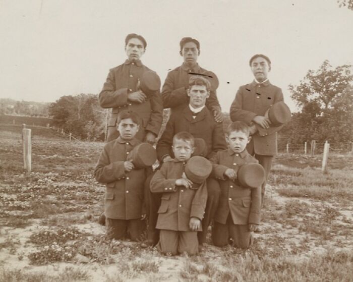 Group portrait of Native American boys and a teacher dressed in early 1900s uniforms at a boarding school.