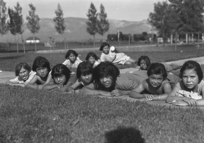 Group of Native American children lying on grass at a boarding school, early 20th century, with distant mountains and trees visible.
